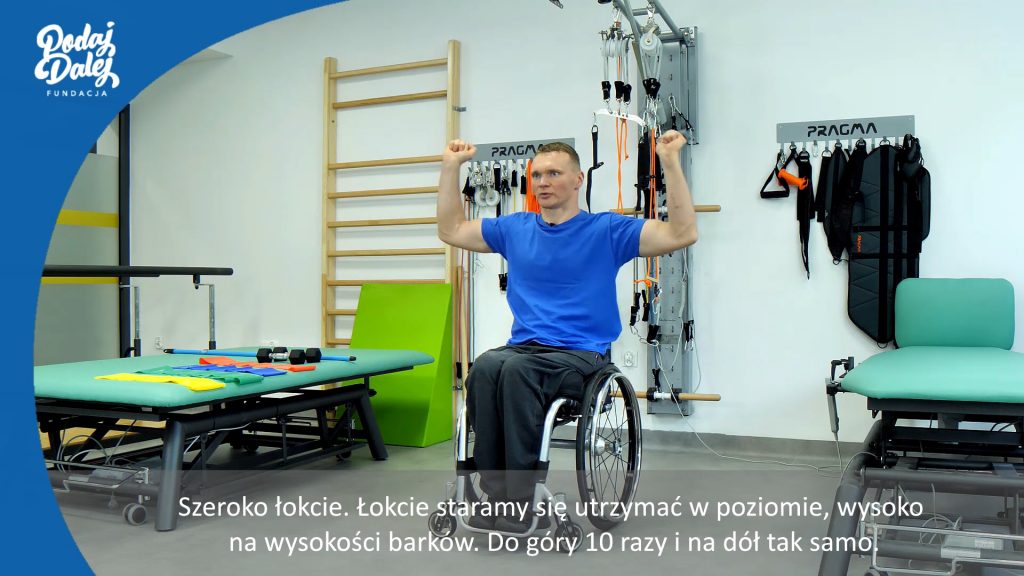 kadr z filmu szkoleniowego, mężczyzna na wózku inwalidzkim unosi ręce do góry. Sala rehabilitacyjna, w tle drabinki, materac.