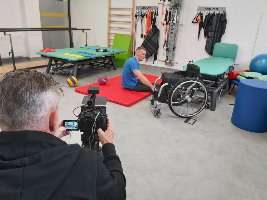 sala gimnastyczna, na materacu siedzi mężczyzna, obok jego wózek inwalidzki