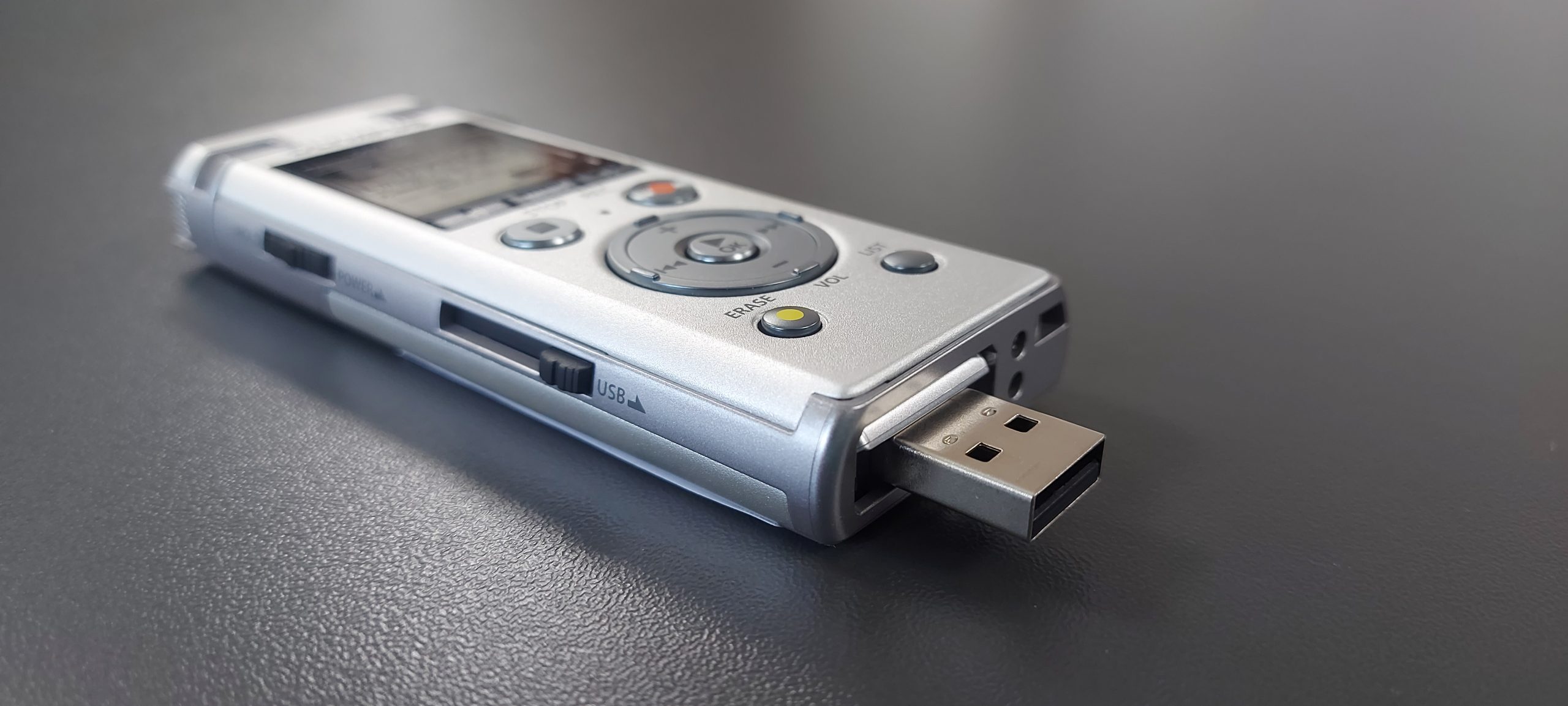 Zdjęcie przedstawia dyktafon Olympus DM-720 z otwartym portem USB, na szarym tle