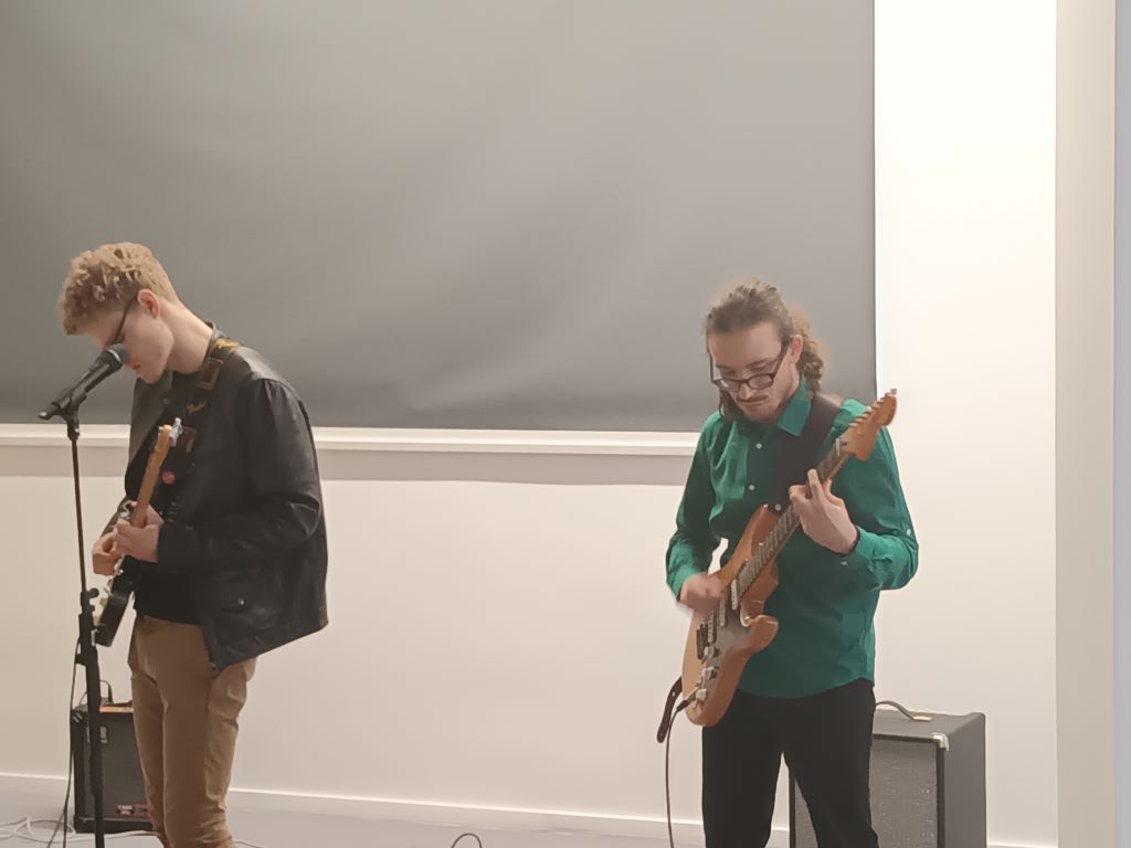 Z lewej strony Łukasz Krasiejko śpiewa i gra na gitarze. Z prawej strony Maksym Bartczak śpiewa i gra na gitarze